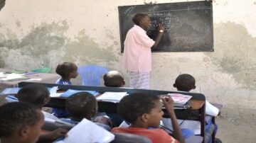 مدرس يشرح درسا على السبورة أمام مجموعة من الطلاب في فصل دراسي بسيط. الطلاب جالسون على مقاعد، يركزون على المعلم ويتفاعلون مع الدروس.