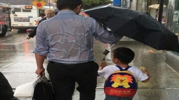 رجل يحمل مظلة بينما يسير على رصيف مبلل بالمطر، وطفل صغير بجانبه يحمل حقيبة ظهر مزينة برسم أسد. في الخلفية، تظهر حافلات ومشاة آخرون في يوم ممطر.