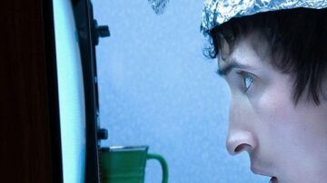 رجل يرتدي قبعة من رقائق الألومنيوم يجلس أمام تلفاز قديم، يظهر على وجهه تعبير مندهش أو مهتم. يركز نظره على الشاشة بينما تضيء الغرفة من ضوء التلفاز.