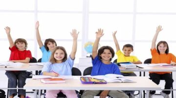 مجموعة من الأطفال في فصل دراسي يرفعون أيديهم للتعبير عن رغبتهم في المشاركة. يرتدون قمصان ملونة ويجلسون على مقاعد أمام مكاتبهم. في الخلفية، توجد خريطة عالمية وأدوات مدرسية، مما يبرز بيئة تعليمية نشطة.