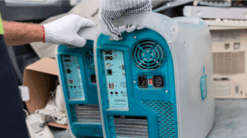 صورة لموظف يرتدي قفازات بيضاء يقوم بفحص خلفية جهاز كمبيوتر قديم، يظهر فيه منافذ توصيل متعددة. يحيط بالجهاز أجهزة إلكترونية قديمة أخرى، مما يشير إلى عملية إعادة تدوير الإلكترونيات.