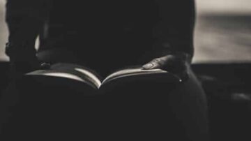 يد تحمل كتابًا مفتوحًا في ظل منخفض، مع التركيز على تفاصيل اليد والصفحات. تعكس الصورة أجواءً من التأمل والهدوء، مما يعزز أهمية القراءة في تعزيز الفكر والإبداع.