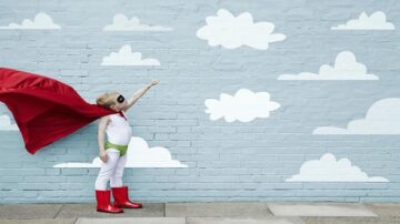 طفل يرتدي زي بطل خارق مع عباءة حمراء وحذاء لامع، يقف أمام جدار مزين بسحب بيضاء، يشير بيده نحو السماء.