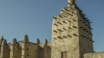 مبنى تاريخي في مالي يظهر عمارة تقليدية مع أسطح مدببة وأعمدة بارزة. السماء صافية وزرقاء، مما يبرز تفاصيل الهيكل المعماري.