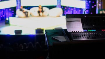 صورة توضح لوحة تحكم صوتية في حدث على المسرح، مع وجود متحدثين يجلسان على الأريكة في الخلفية. الإضاءة الملونة والخلفية المزينة تضيف جوًا احتفاليًا. تحتوي الصورة على تفاصيل واضحة لمعدات الصوت، مما يبرز التنظيم والتقنية المستخدمة في الفعاليات.