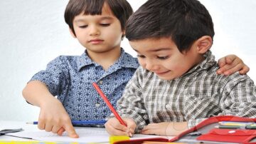 طفلان يجلسان على طاولة، أحدهما يشير إلى ورقة بينما الآخر يكتب بقلم رصاص. يبدو عليهما التركيز والسعادة أثناء التعلم.