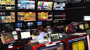 غرفة أخبار تلفزيونية تحتوي على شاشات عرض متعددة تعرض تغطيات إخبارية متنوعة، مع وجود موظفين يعملون على أجهزة الكمبيوتر. تُظهر البيئة تركيزًا على تكنولوجيا البث وإنتاج الأخبار.