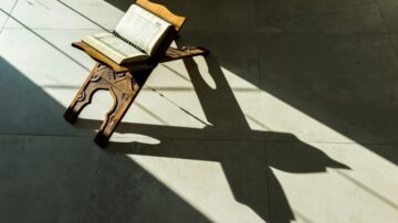 كتاب مفتوح على حامل خشبي، تسقط ظلاله على الأرض. الإضاءة الطبيعية تبرز تفاصيل الكتاب والخشب.