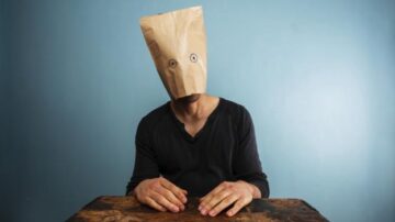 شخص يجلس على طاولة مع كيس ورقي على رأسه، يظهر عينيه مرسومتين على الكيس، والخلفية زرقاء. تعبير الوجه غير مرئي بسبب الكيس الورقي.