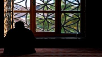 شخص جالس في زاوية غرفة مظلمة، يواجه نافذة مزخرفة بأشكال هندسية. الضوء الطبيعي يتسلل عبر النافذة، مما يخلق تباينًا بين الظل والضوء. تعكس الألوان الخضراء خارج النافذة طبيعة هادئة.