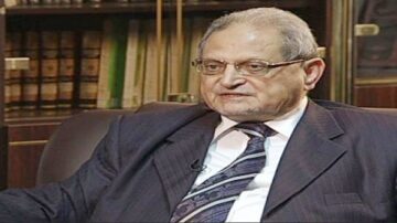 رجل مسن يرتدي بدلة في غرفة بخلفية رفوف كتب.