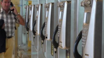 صورة لصف من الهواتف العمومية المعدنية في مكان عام، مع وجود رجل يتحدث في أحد الهواتف. تعكس الصورة مشهداً من الحياة اليومية في المناطق الحضرية، مما يبرز استخدام التكنولوجيا القديمة في الاتصال.