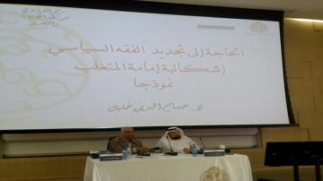 صورة لمؤتمر يتناول موضوع الحاجة لتجديد الفقه السياسي، حيث يظهر متحدثان أمام شاشة تعرض عنوان الجلسة. الخلفية تحمل شعارات المؤسسة المنظمة، بينما يركز الحضور على المناقشة.