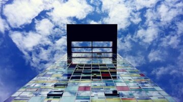 صورة عمارة حديثة بواجهة زجاجية ملونة تعكس السماء الزرقاء والغيوم. تُظهر الصورة زاوية منخفضة للبرج مع إطار أسود في الأعلى، مما يبرز التصميم المعماري الفريد.