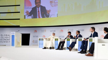 مؤتمر القمة العالمية للاقتصاد الإسلامي، يتضمن مجموعة من المتحدثين على منصة، يشاركون في مناقشة حول الاقتصاد الإسلامي. يظهر في الصورة متحدث رئيسي يتحدث بينما يجلس أربعة آخرون على المنصة. الخلفية تحمل شعار القمة ومدينة حديثة.