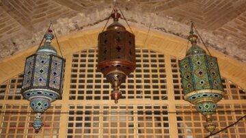 ثلاثة فوانيس ملونة معلقة تحت قوس، مع تفاصيل مزخرفة، تعكس التصميمات التقليدية في العمارة الإسلامية. الفوانيس تحتوي على ألوان زاهية وأنماط هندسية، مما يضفي جوًا دافئًا ومميزًا للمكان.