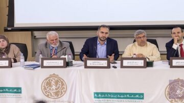 ندوة في جامعة حمد بن خليفة، تتضمن خمسة متحدثين يجلسون خلف طاولة، مع وجود لافتات للجامعة. المتحدث الرئيسي يرتدي بدلة ويشير بيده، بينما الآخرون يستمعون بتركيز. تظهر خلفهم لافتة تحمل شعار الجامعة.