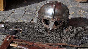 خوذة حديدية تاريخية مع درع من حلقات معدنية وسيف موضوعين على أرضية حجرية.