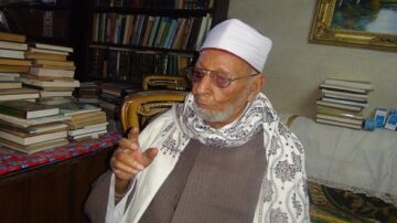 رجل مسن يرتدي عمامة بيضاء ونظارات، يجلس في غرفة مليئة بالكتب. يظهر خلفه رفوف الكتب ومكتب بسيط، مع لمسات من الديكور التقليدي. تعكس تعابير وجهه الحكمة والخبرة.