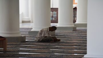 رجل يصلي في مسجد، يجلس على الأرض بين أعمدة بيضاء وسجادة مزخرفة، يركز على عبادته في بيئة هادئة.