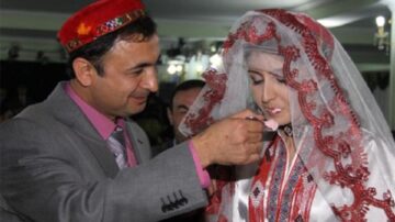 رجل يرتدي قبعة تقليدية يقدم لقمة طعام لامرأة ترتدي فستان زفاف مزين بتطريزات تقليدية وحجاب. المشهد يعكس تقاليد الزفاف في الثقافة.