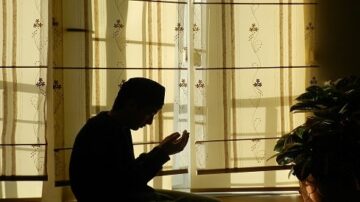 شخص يجلس في غرفة مضاءة بأشعة الشمس، يظهر ظله خلف ستائر مزخرفة. يضع يديه في وضع دعاء، مع التركيز على التأمل والسكينة.