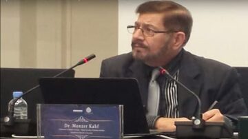 دكتور مؤثر كاف يجلس أمام حاسوب محمول خلال مؤتمر، يتحدث بلطف مع وجود ميكروفونات على الطاولة. خلفه لافتة تحمل اسمه، مع زجاجة ماء بجانبه.