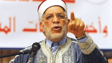 شخص يرتدي لباس تقليدي يتحدث بحماس في مؤتمر، يشير بيده نحو الجمهور، مع وجود لافتة خلفه.