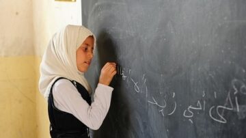 طفلة ترتدي الحجاب تكتب على سبورة سوداء في فصل دراسي. تركز على الكتابة بأسلوب دقيق وتعبر عن اهتمامها بالتعلم.