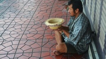 رجل يجلس على الرصيف في منطقة حضرية، يحمل سلة في يديه. يرتدي قميصًا غير مرتب ويبدو عليه التعب. الأرضية مصنوعة من بلاط ملون بأشكال هندسية.