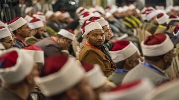 رجال يرتدون عمامات حمراء يجلسون في قاعة، مع التركيز على رجل يلتفت نحو الكاميرا. الجو يعكس تجمعًا دينيًا أو ثقافيًا.