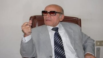 رجل مسن يرتدي نظارات شمسية، يجلس على كرسي في مكتب. يرتدي بدلة رمادية وربطة عنق. يضع يده في وضع تعبير عن الحديث. خلفية بسيطة.