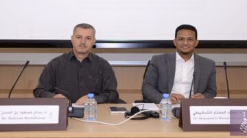 صورة لرجلين يجلسان على طاولة في قاعة مؤتمرات. الرجل على اليسار يرتدي قميصًا أسود ويظهر اسم الدكتور بلقاسم بن عيسى على اللوحة أمامه. الرجل على اليمين يرتدي قميصًا فاتحًا ويظهر اسم الدكتور محمد الحسن المنصوري. يوجد زجاجة ماء وميكروفون على الطاولة.