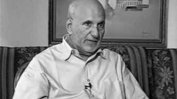 رجل مسن ذو شعر أصلع يجلس في غرفة مع أريكة مزخرفة. يرتدي قميصًا فاتح اللون، ويظهر تعبيره جادًا. خلفه لوحة تحتوي على معالم معمارية.