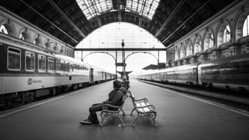 رجل يجلس على مقعد في محطة قطار مهجورة، مع قطارين في كلا الجانبين. السقف الزجاجي الكبير يخلق جوًا دراماتيكيًا، بينما تعكس الألوان الرمادية مشاعر الهدوء والانتظار.