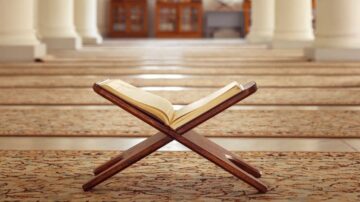 كتاب مفتوح على حامل خشبي في مسجد، مع تفاصيل الأرضية المزخرفة والأعمدة الخلفية، يرمز إلى العبادة والتأمل.