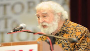 رجل مسن ذو لحية وشعر أبيض يتحدث من خلف ميكروفون على منصة، يرتدي قميصًا مزخرفًا بألوان دافئة. يظهر خلفه لافتة تشير إلى حدث تاريخي أو ثقافي.