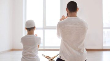 رجل وطفل يجلسان معًا في غرفة مضاءة بشكل جيد، يتجهان نحو مصحف موضوع أمامهما. كلاهما في وضع صلاة، مع أيديهم مرفوعة للدعاء. الأجواء هادئة وتعكس روحانية اللحظة.