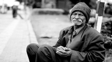 رجل مسن يجلس على الرصيف في مدينة، يرتدي سترة داكنة وقبعة، ويبدو عليه علامات التعب. تعكس تعابير وجهه الصعوبات التي يواجهها.