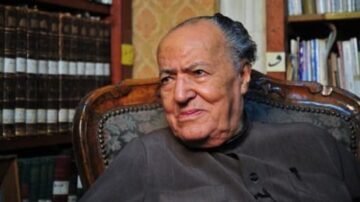 رجل مسن يجلس على كرسي مزخرف في مكتبة تحتوي على مجموعة من الكتب. يظهر تعبيره الودود وهو يرتدي ملابس تقليدية. الخلفية تضم رفوف كتب قديمة، مما يبرز أجواء ثقافية وعلمية.