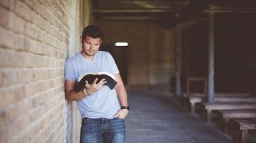 رجل شاب يرتدي قميصاً رمادياً ويقرأ كتاباً بينما يستند إلى جدار من الطوب في مكان داخلي، مع مقاعد خشبية في الخلفية.