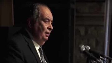 رجل يتحدث أمام ميكروفونات في حدث رسمي، يرتدي بدلة سوداء، يظهر التركيز والجدية.