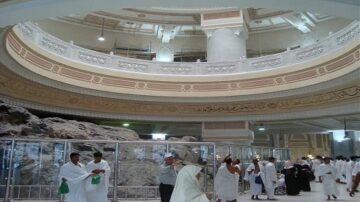 صورة لزوار يطوفون حول الكعبة في مكة المكرمة داخل المسجد الحرام. يظهر التصميم المعماري الفاخر للمسجد، مع تفاصيل زخرفية وكتابات عربية. الحجاج يرتدون ملابس الإحرام البيضاء، ويستمتع البعض بأجواء الروحانية.