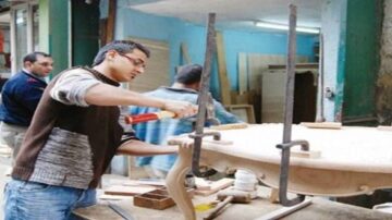 شاب يعمل في ورشة نجارة، يستخدم أدوات لتجميع قطعة أثاث خشبية. يظهر في الخلفية عدد من العمال في بيئة صناعية.