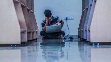 شاب يجلس على الأرض في ممر ضيق، يعمل على اللابتوب. يظهر التركيز على وجهه، مع وجود كابلات كهرباء بالقرب منه. ينعكس الضوء على الأرضية اللامعة.