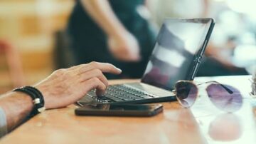 يد تظهر يد شخص يستخدم جهاز كمبيوتر محمول على طاولة، مع نظارات شمسية وهاتف ذكي بجانبه. تعكس الصورة بيئة عمل غير رسمية تعزز من استخدام التكنولوجيا في الحياة اليومية.