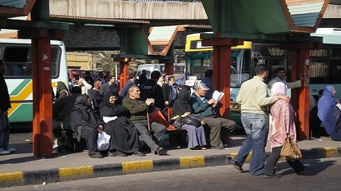 موقف حافلات مزدحم في مدينة بها أشخاص ينتظرون، يجلسون، يقفون، ويمشون.