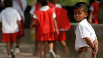 طفل يرتدي زي المدرسة الأبيض مع بنطال بني، ينظر إلى الكاميرا بينما يتجه زملاؤه في الخلفية نحو منطقة أخرى، يرتدون زيًا أحمر. تعكس الصورة لحظة من الحياة المدرسية وتفاعل الأطفال في بيئة تعليمية.