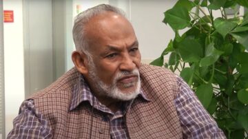 رجل مسن ذو لحية رمادية وشعر أبيض، يرتدي سترة مزخرفة وقميص مربع، يجلس في مكان داخلي مع نباتات خضراء خلفه. يبدو على وجهه تعبير جاد وهو ينظر إلى الأمام.