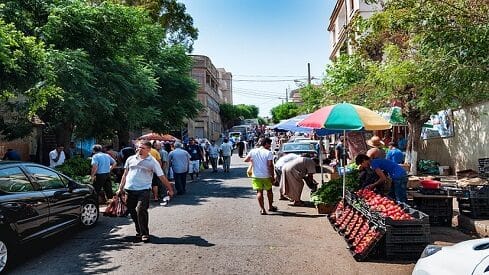 سوق مفتوح في شارع تتجول فيه الناس لشراء الخضروات والفواكه بالقرب من المباني والسيارات.